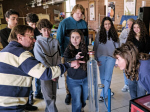 Un groupe de jeunes, accompagné d’un animateur et d’une animatrice, observe une expérience scientifique autour d’un grand tube en verre rempli de liquide, dans une salle de physique remplie de matériel éducatif.
